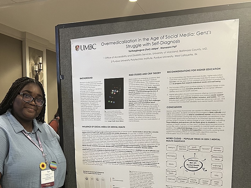A smiling  woman with long, braided hair stands to the left of a large, scientific poster board. The poster is titled "Overmedicalization in the Age of Social Media: GenZ's Struggle with Self-Diagnosis." The poster has sections including "Background," "Bad Studies and Crip Theory," "Recommendations for Higher Education," and "Conclusion." The woman is wearing a light blue, collared shirt and dark pants. A conference lanyard with a nametag hangs around her neck, and a sunflower pin is attached to the lanyard. She is looking directly at the camera. In front of the poster board on a covered table, there is a small cup of water, a yellow sticky note, and some other small objects. The poster board itself is on an easel. The background appears to be a conference hall with beige walls and a lamp on the far left side of the image.