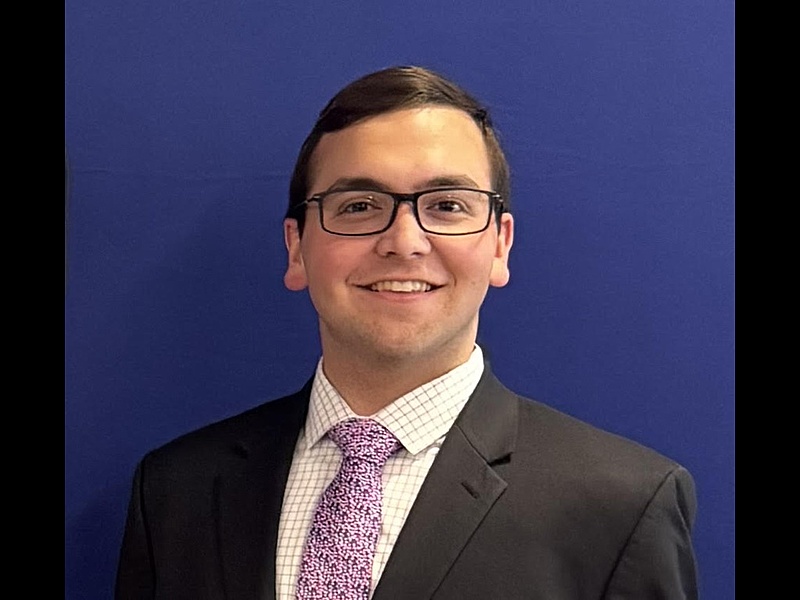 paul in a suit and tie standing in front of a blue background smiling