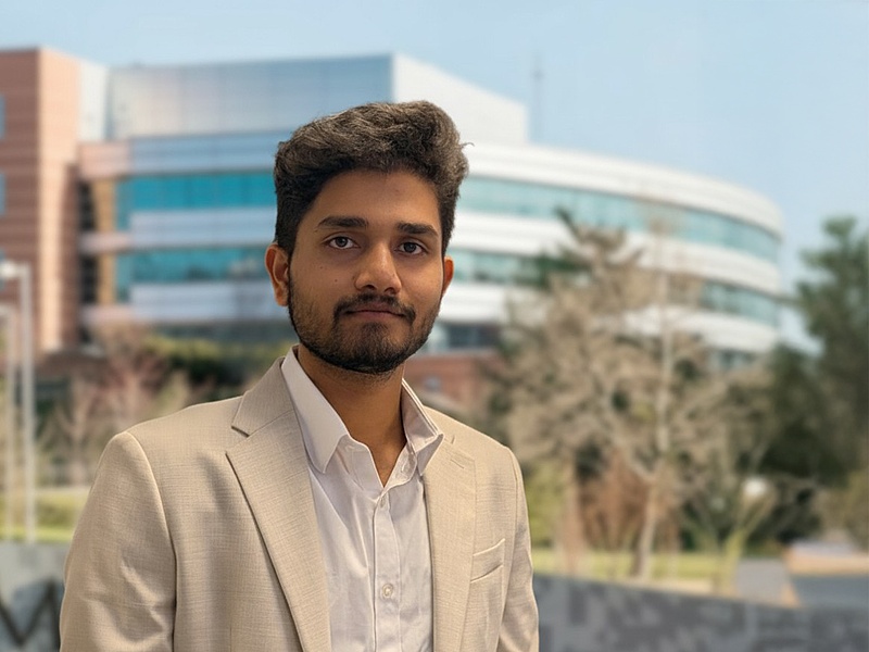 human with mustache and beard wearing a while shirt and tan jacket, standing in front of a building.