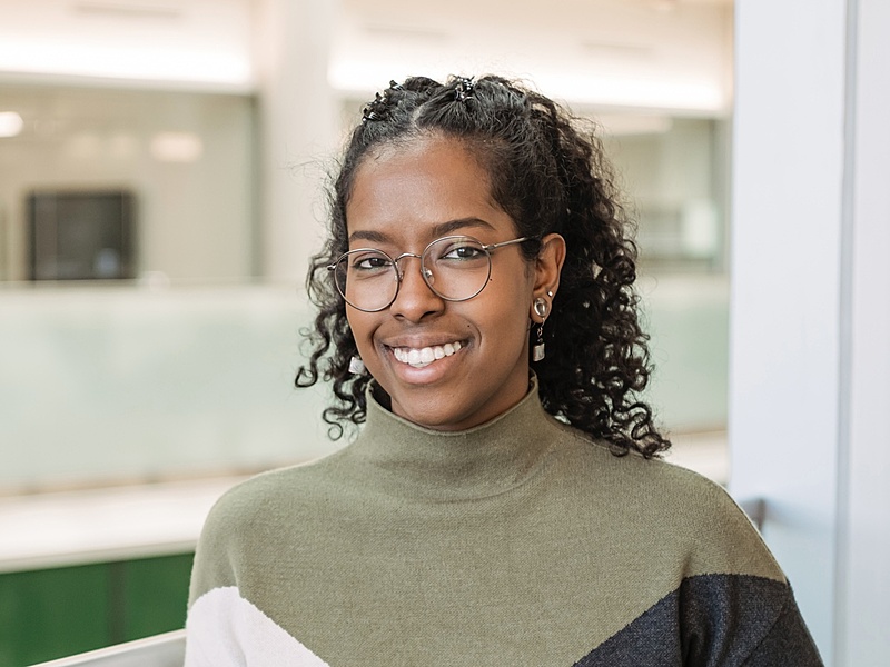 A smiling woman with curly hair pulled half back stands indoors near a glass railing. She wears round glasses, small drop earrings, and a color-block turtleneck sweater in olive green, cream, and dark gray. The background is softly blurred, showing a bright, modern interior space.