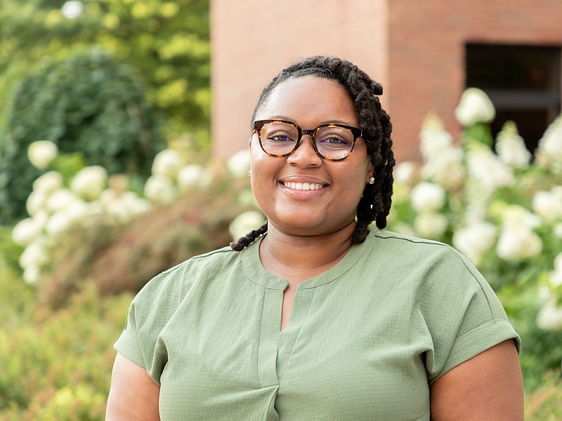 human with short hair in a green shirt standing in front of a garden and building