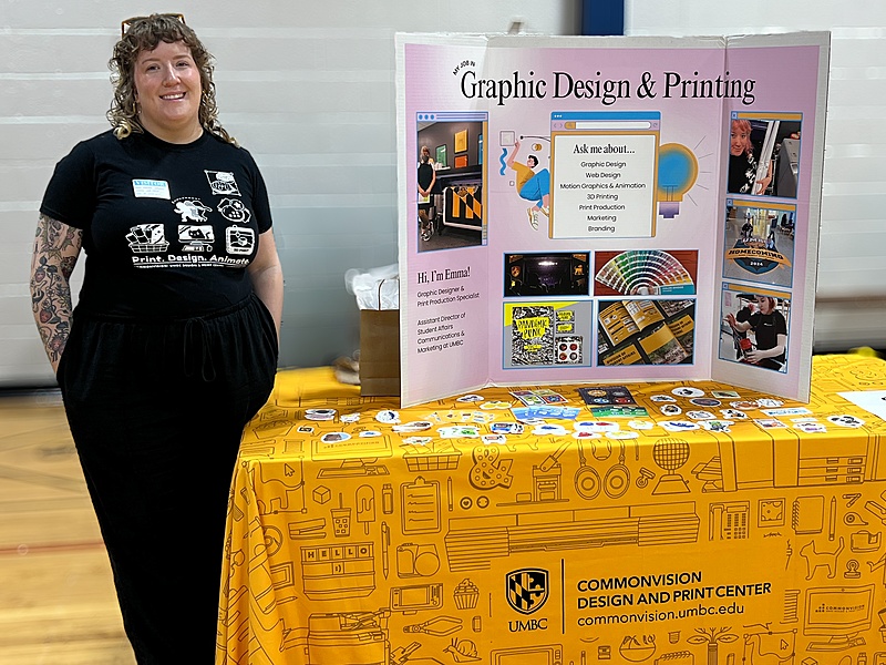 Human with shoulder length hair standing next to a poster on a table.