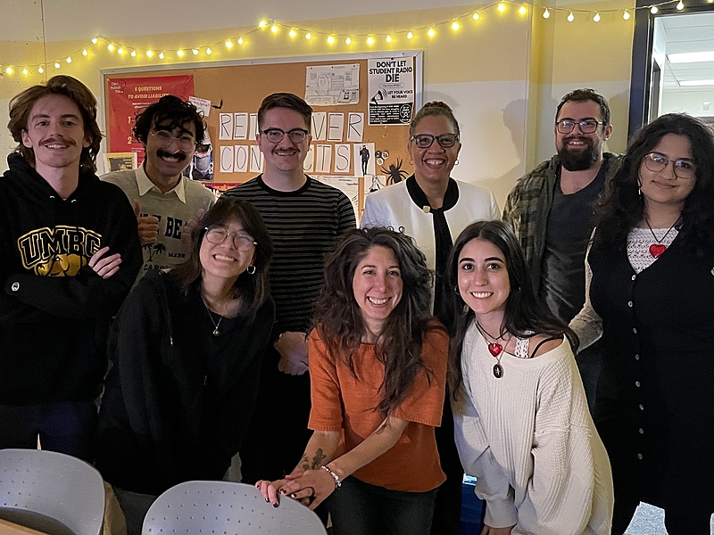 A group of humans in an office in front of a bulletin board and string lights.