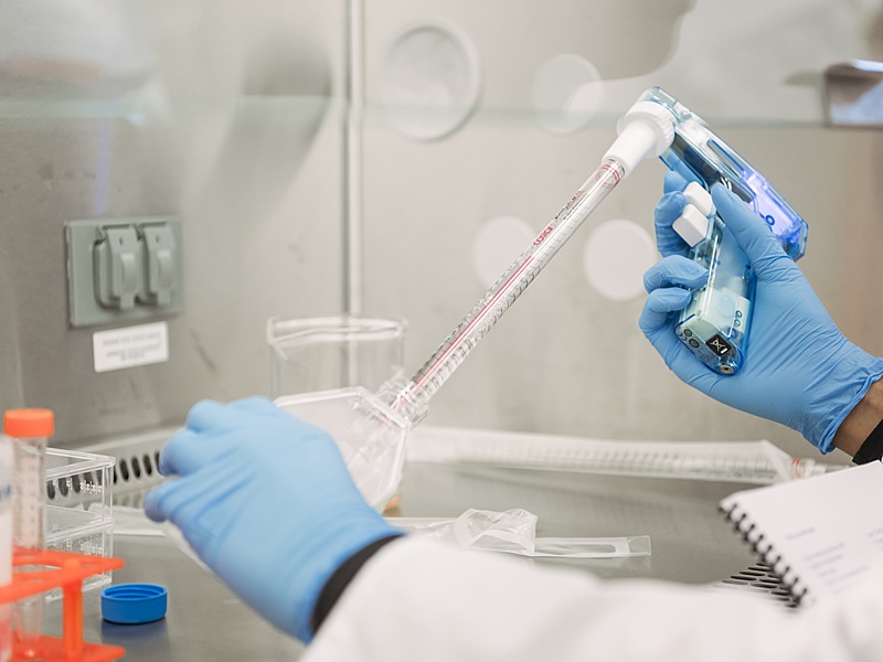 A pair of gloved hands, wearing light blue nitrile gloves, are working in a laboratory setting. The person on the left is holding a large, clear, plastic vessel with a conical bottom, which is attached to a long, red-and-white striped pipette. The pipette is connected to a white hand-held device. The person's other hand, on the right, is holding a smaller clear bottle with a blue liquid inside, which is also attached to a pipette-like device. The bottle appears to be glowing blue. The background shows a stainless steel fume hood or biosafety cabinet. There are other lab items on the counter, including clear plastic tubes in a rack, a white notebook, and an orange-capped bottle. The overall impression is one of scientific research or experimentation.