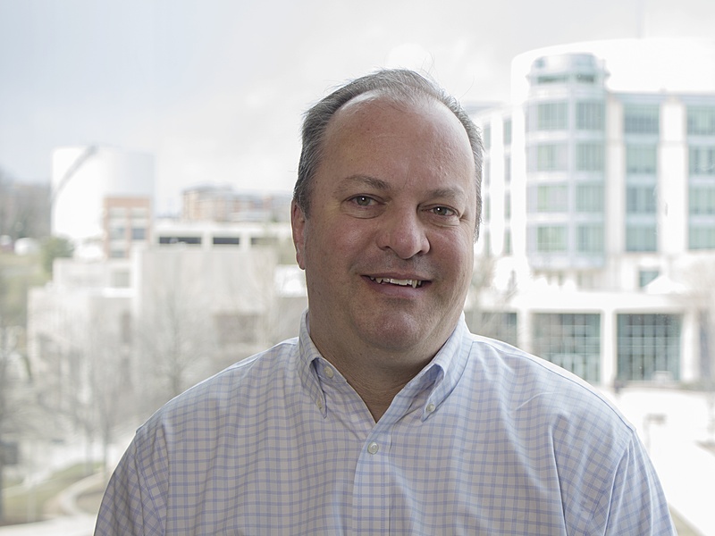 Human with short hair and a collared shirt standing in front of a building.