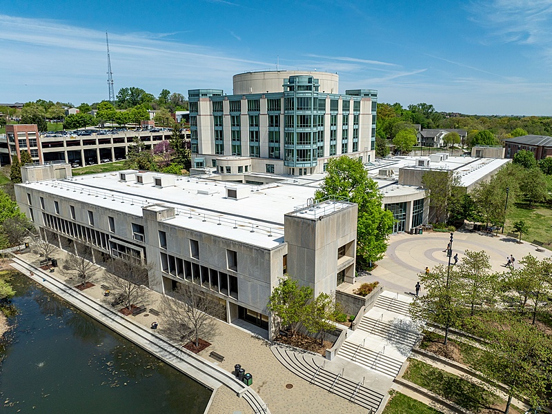 Aerial view of the Albin O. Kuhn Library and Gallery, a modern building with a large circular tower and adjacent rectangular structures. The building is surrounded by green trees and a small pond, under a clear blue sky. A parking garage is located behind the library building.