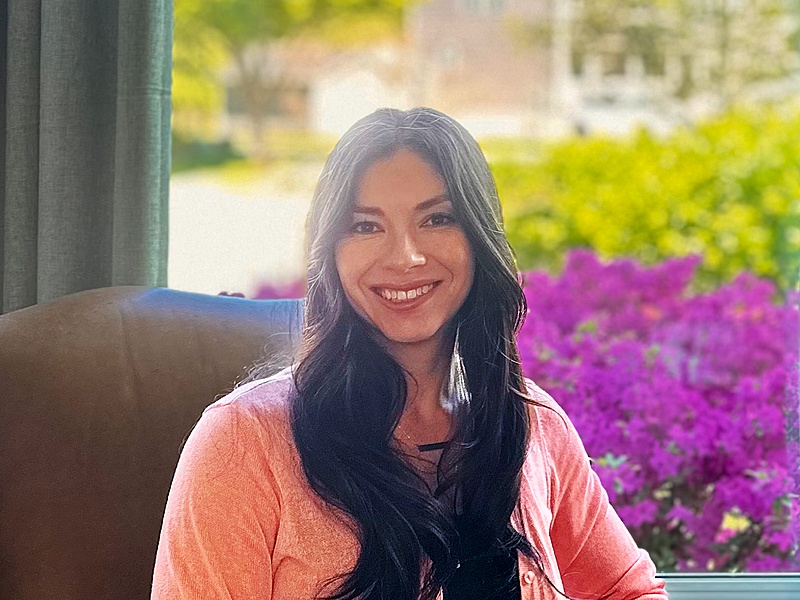 Picture of Dr. Jackson Soller smiling, sat in a chair, with a background of green leaves, trees, a blurred house and purple flowers.