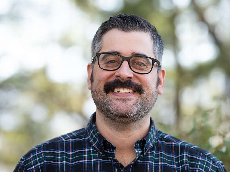 A human wearing glasses and a blue plaid shirt standing in front of trees.