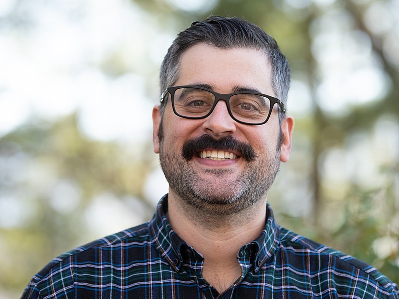 Portrait of Matt Hoffman with short brown hair and glasses wearing a blue plaid shirt, with trees behind him