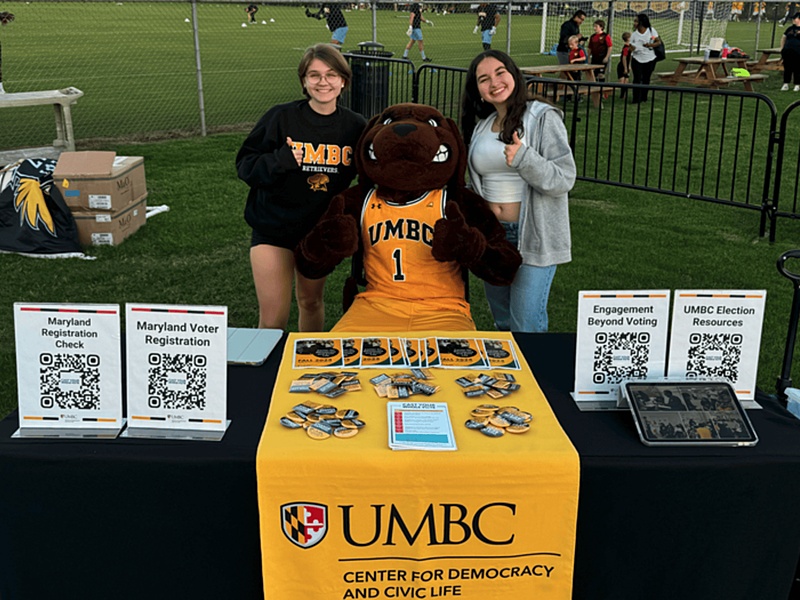 Two Center for Democracy and Civic Life staff members sit with the True Grit mascot and smile at a voter registration and election information table.