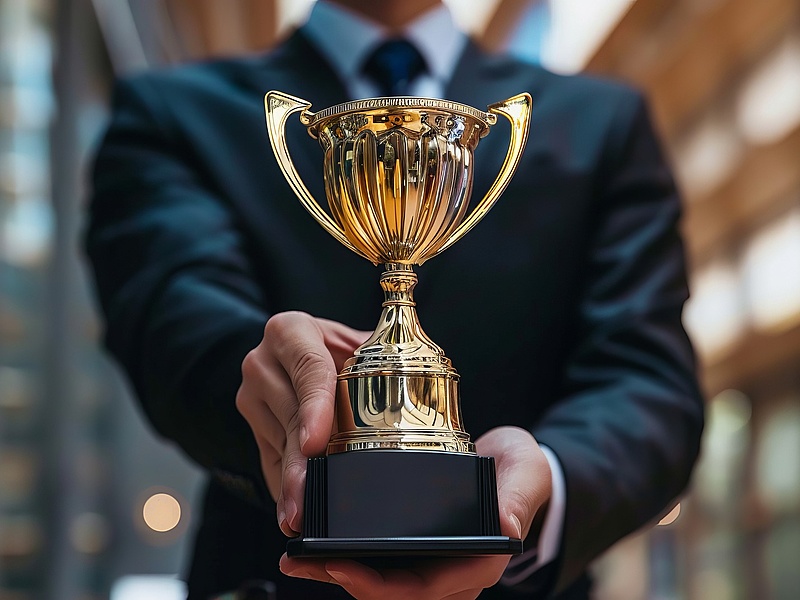 Person in a dark business suit holding a gold trophy with both hands, presenting it forward in a modern indoor setting