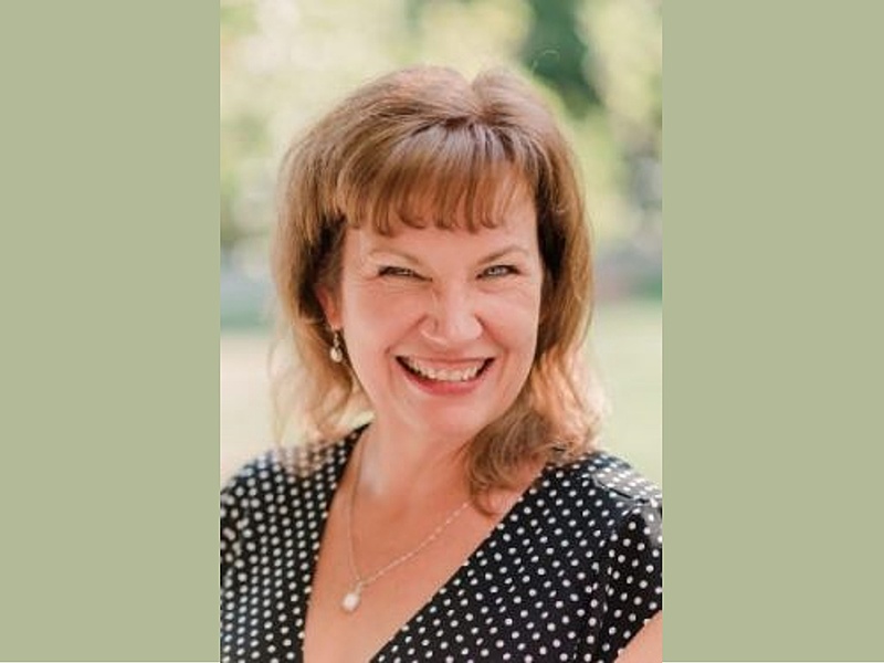 A headshot of a white woman with light brown hair smiling and wearing a black polka-dot shirt and a white pendant necklace.