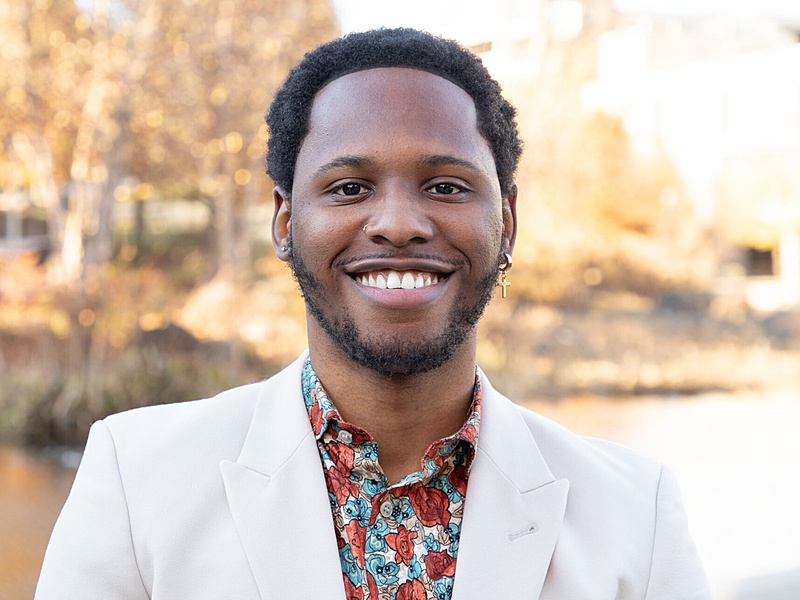 A human wearing a patterned shirt and white jacket standing in front of a pond and trees.