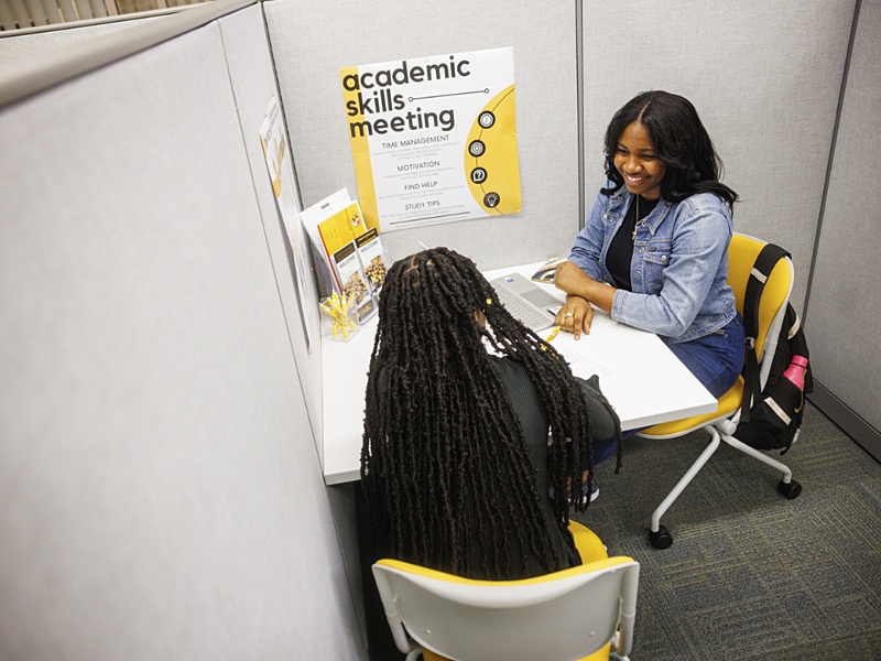 Two students sitting together, having a meeting at a white table/ A poster with the words, "Academic Skills Meeting" is posted on the wall behind them.