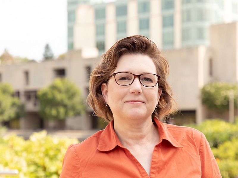 A human with chin length hair and glasses in a orange shirt standing in front of a building and trees.