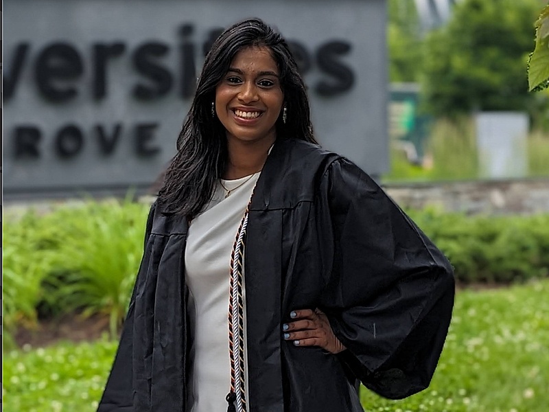 Asmiling graduate wearing a black cap and gown standing outdoors on campus.