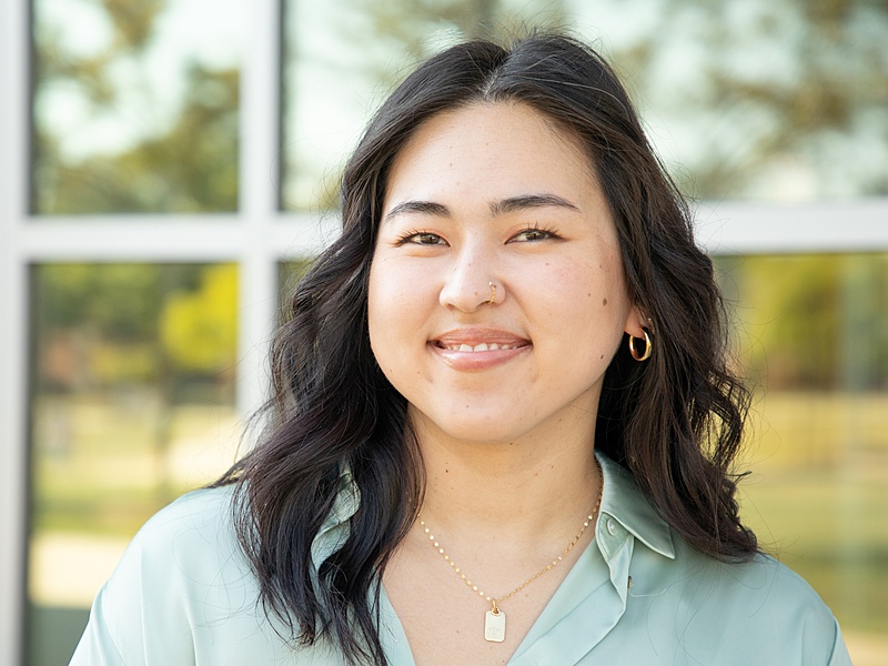Portrait of Angelina Jenkins with shoulder length dark hair, with a nose ring, hoop earrings and a gold necklace, wearing an aqua shirt.