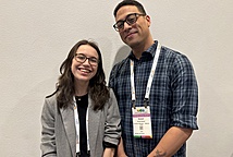 Two smiling humans one with glasses and shoulder length dark hair wearing a black shirt and grey blazer, and one with glasses and short dark hair wearing a lanyard and blue plaid shirt