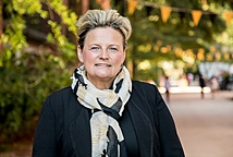 A person with short blonde hair wearing a dark blue blazer and shirt and yellow, white, and dark blue scarf standing in front of trees with green leaves