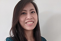 Portrait of Asian American woman with long dark brown hair in a green dress shirt smiling at the camera with a light beige wall in the background