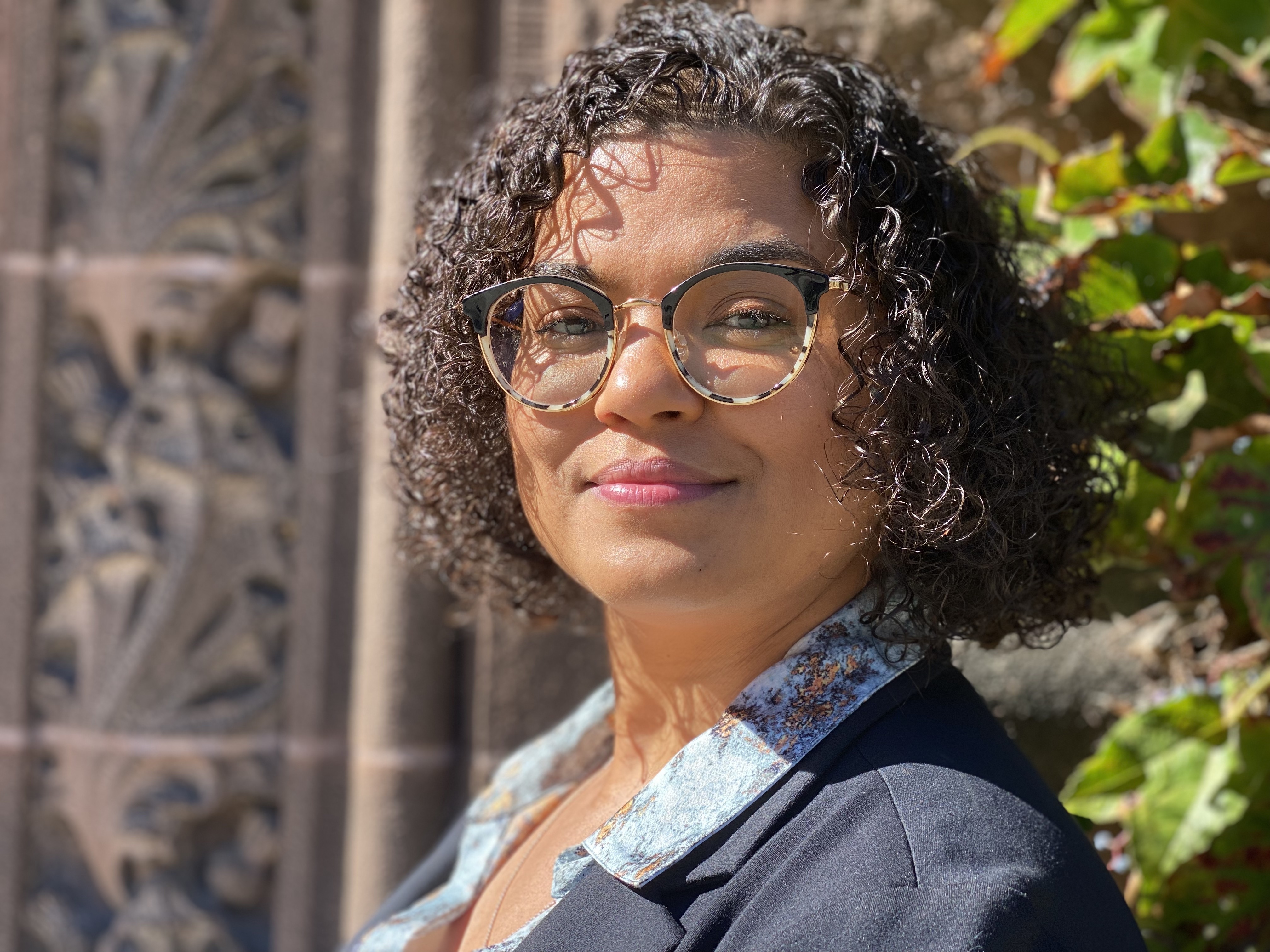woman  with glasses and dark, curly hair, wearing a blue shirt and jacket posing outside