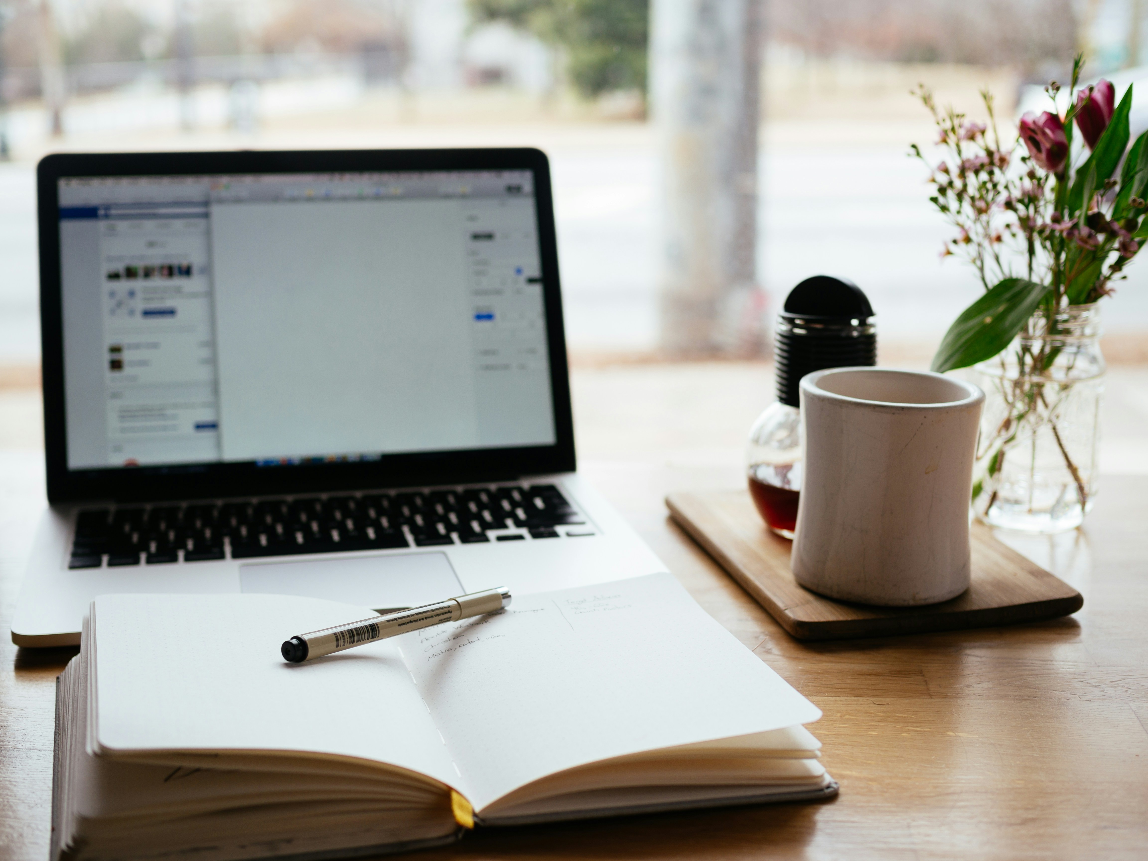 A laptop, notebook, pen, coffee mug, and flowers on a desk
