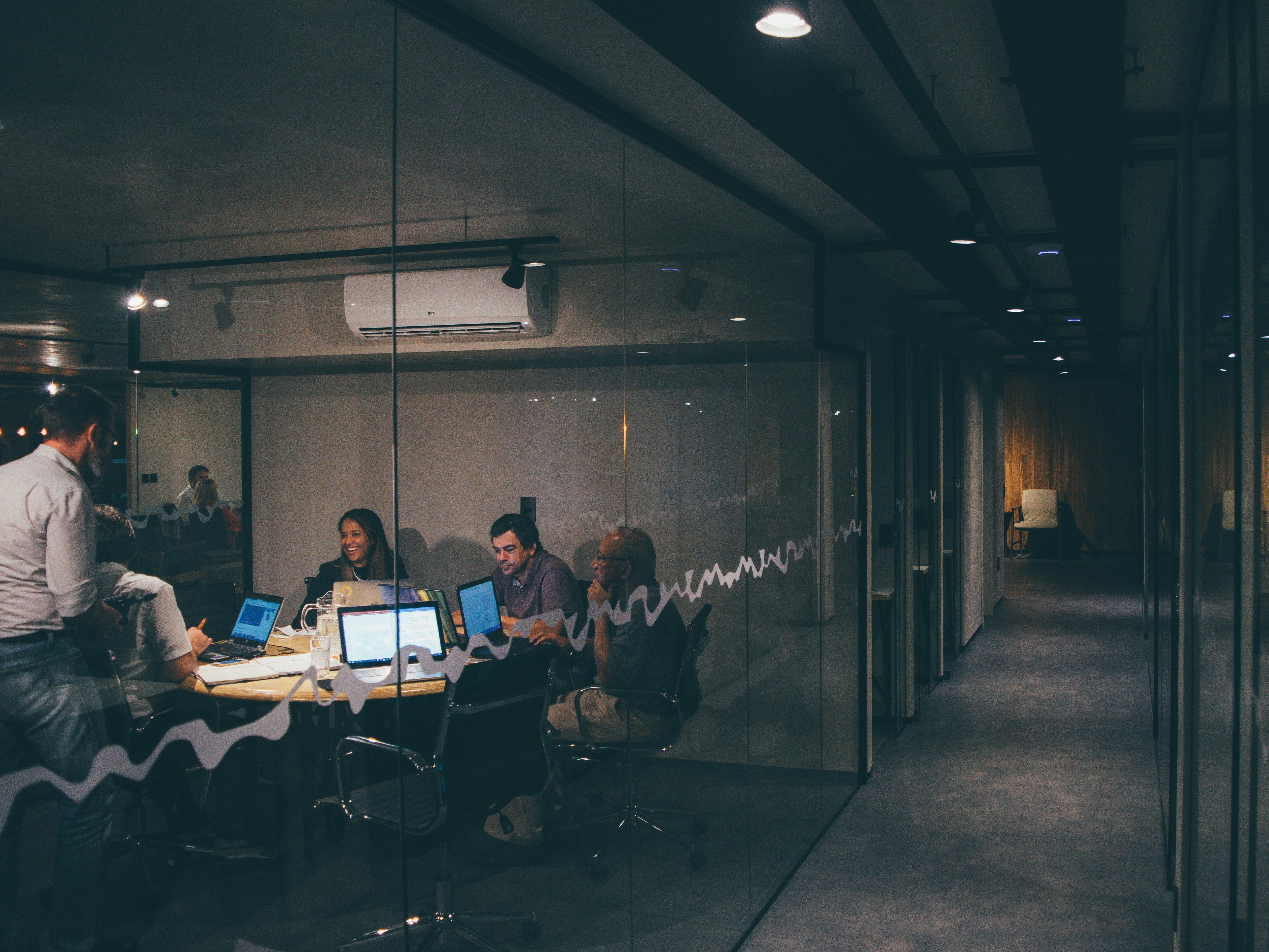 a meeting room with a mix of professionals from different backgrounds, age and genders sit down with their laptops, talking.