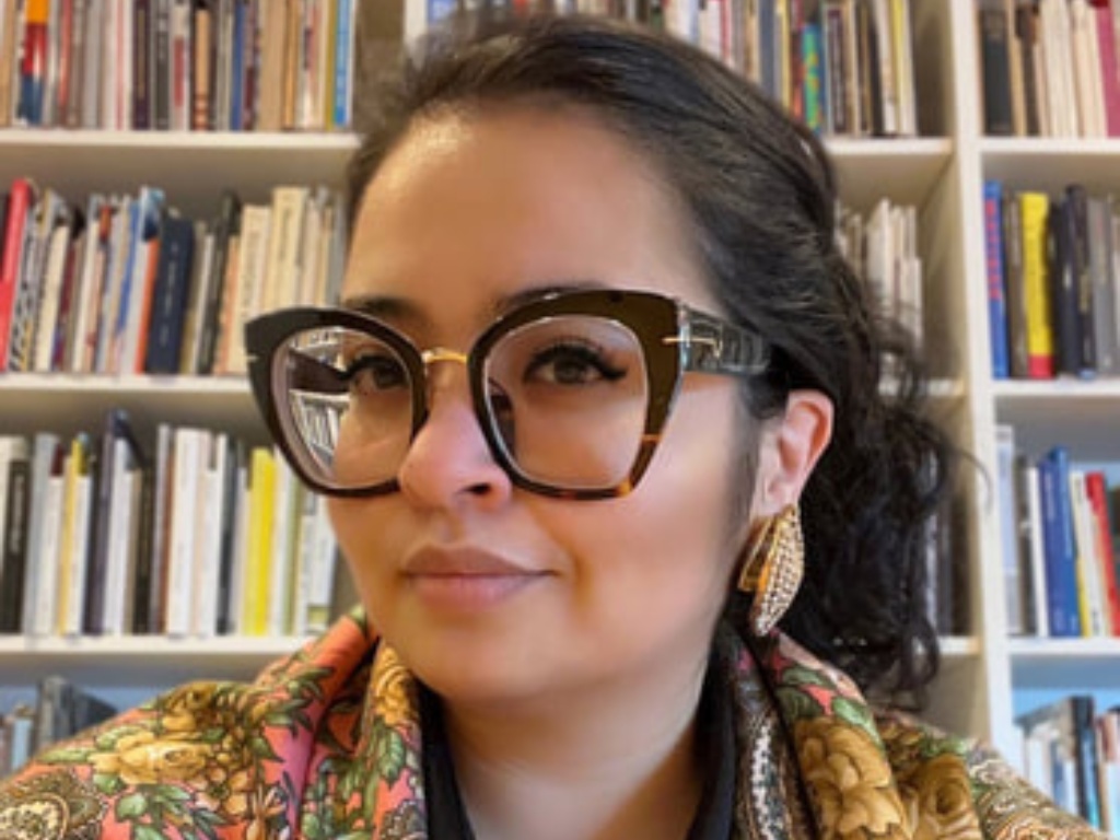 An indigenous women wearing large-framed glasses is smiling while seated in front of a case full of books.