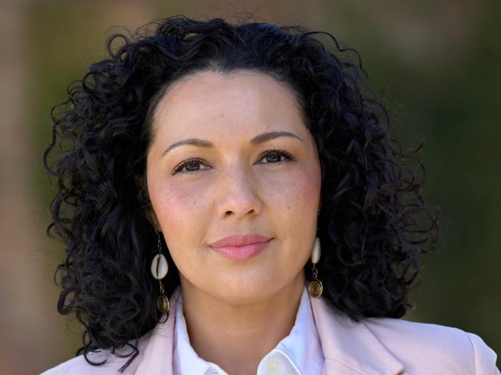 An Afro-Latinx woman with dark curly hair and a pink blazer is smiling.