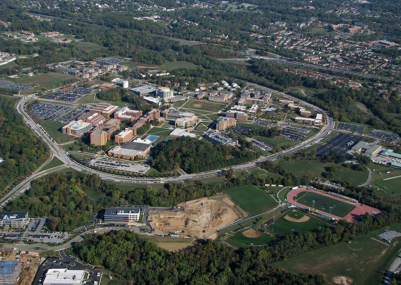 Ariel photo of UMBC's main campus, both academic and residential buildings, within Hilltop Circle. The Research Park and Athletic Fields are in the foreground