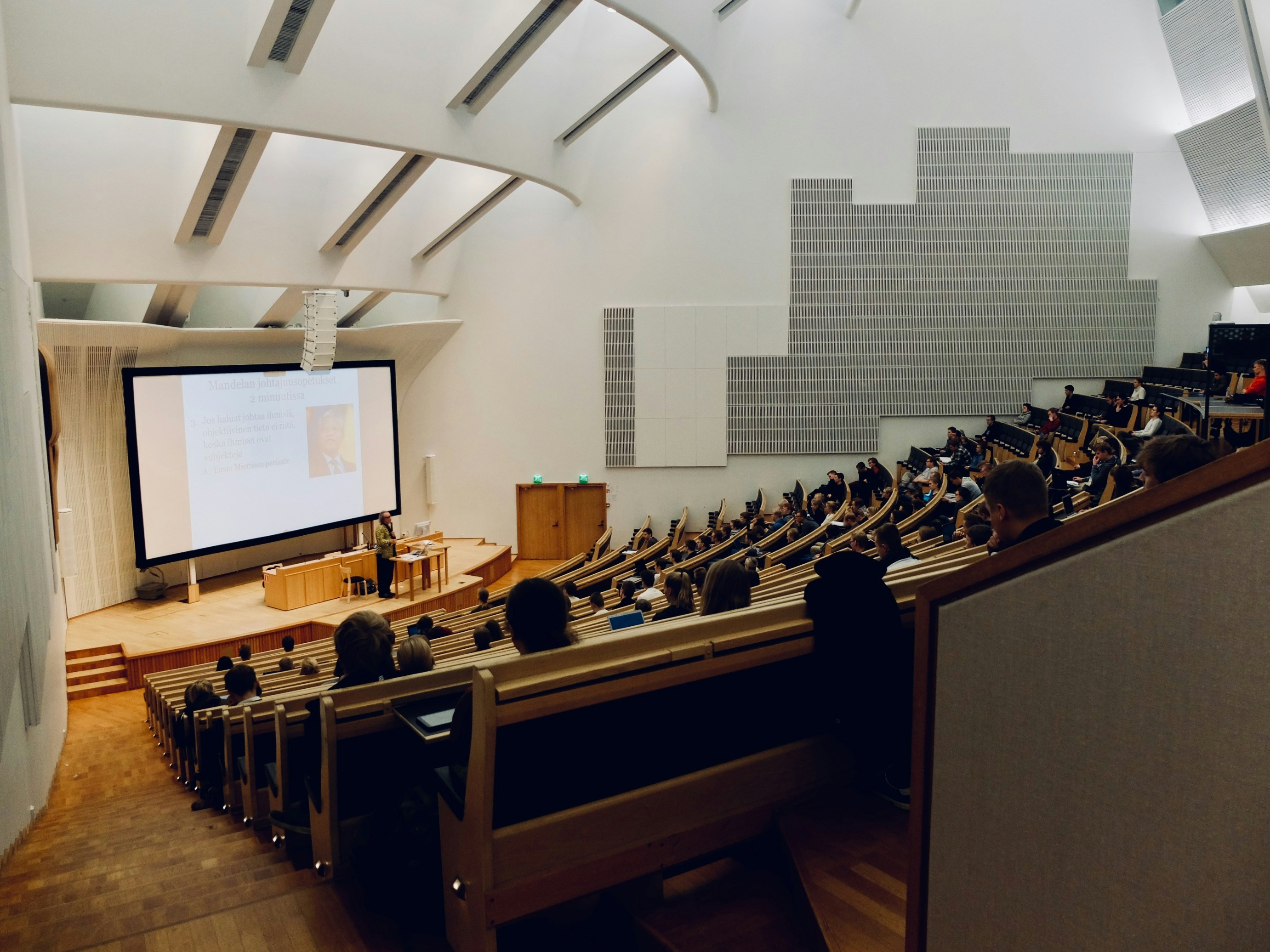 The image depicts a sloping theater style classroom full of students for a presentation.