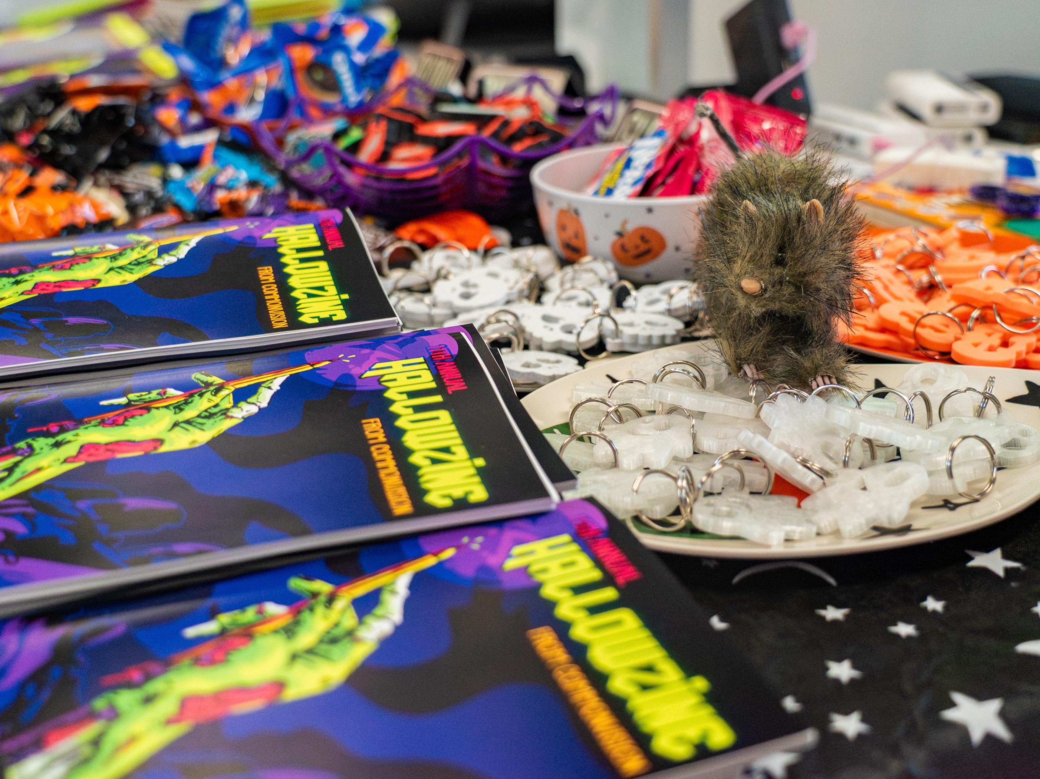 Table full of 2025 Hallowzines, candy, and spooky giveaways. A stuffed rat used for decor checks out the goodies.
