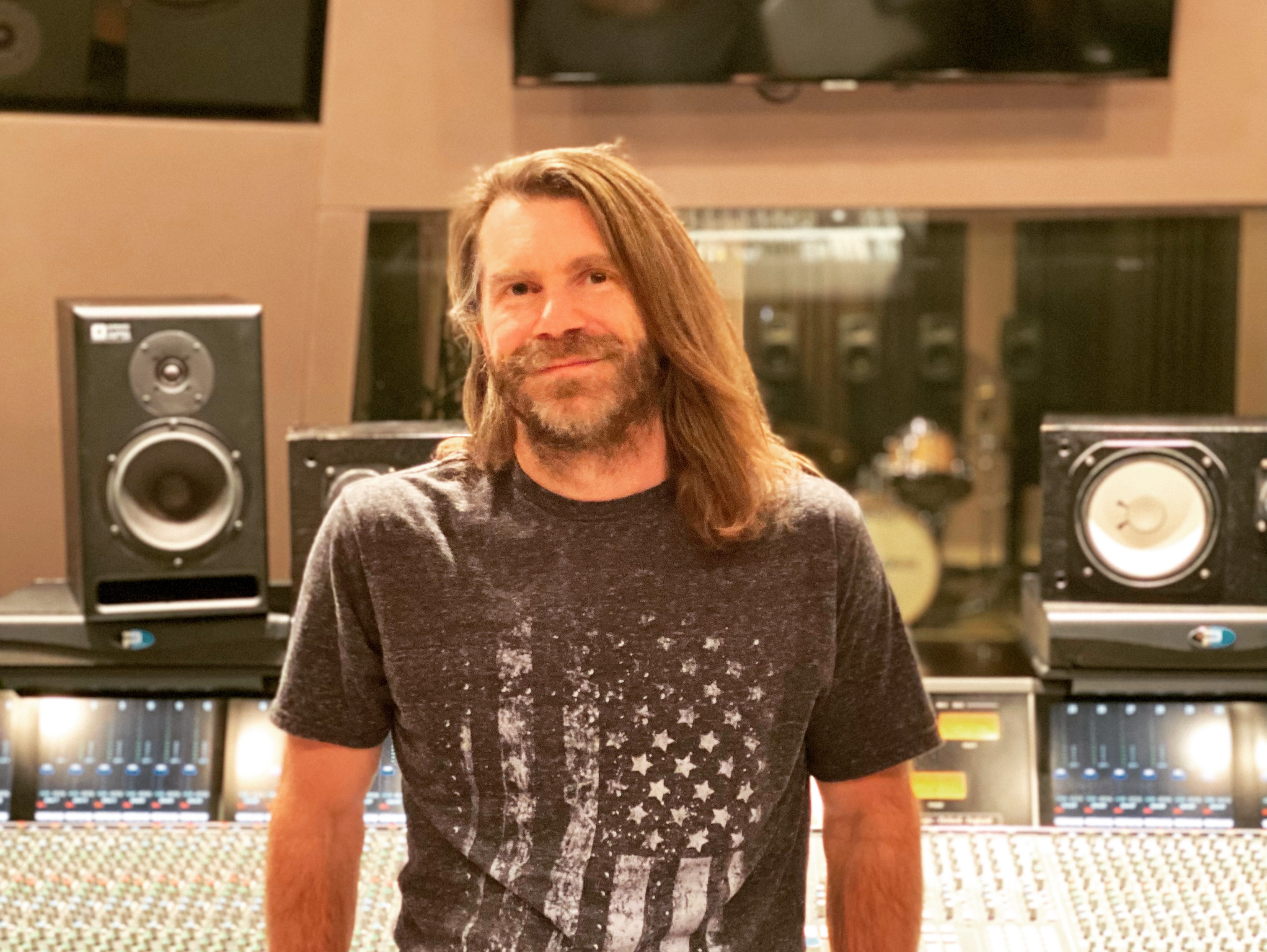 a lighter skinned man with strawberry blonde hair and a beard does a soft smile while sitting on a digital mixing booth.