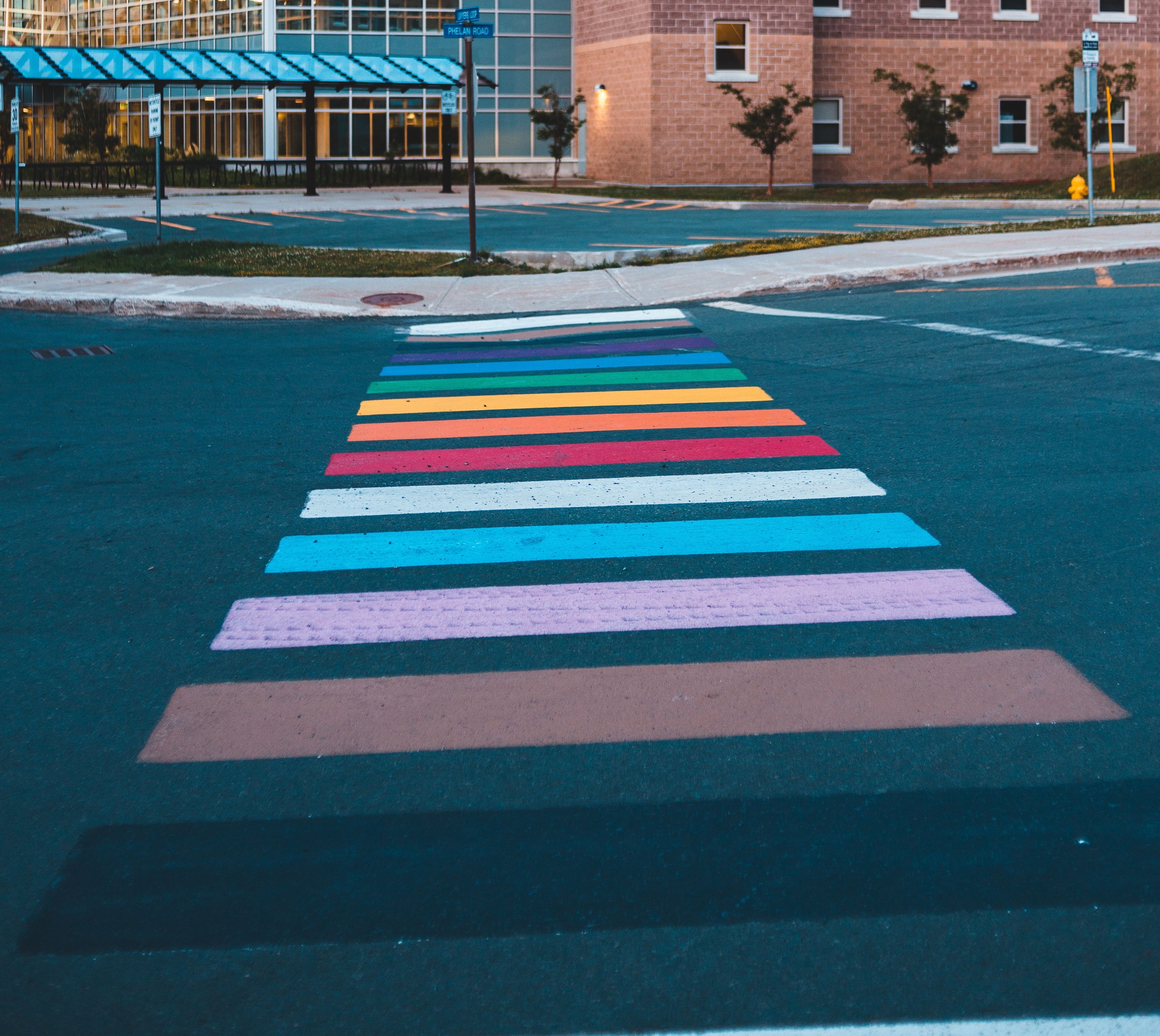 a crosswalk to a curb cut ramp in Progress Pride colors - black, brown, pastel and rainbow stripes mark the path