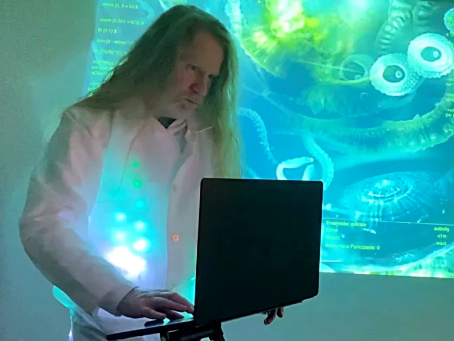 a long haired person stands in front of a sea-like blue and green projection screen and does something on a computer.