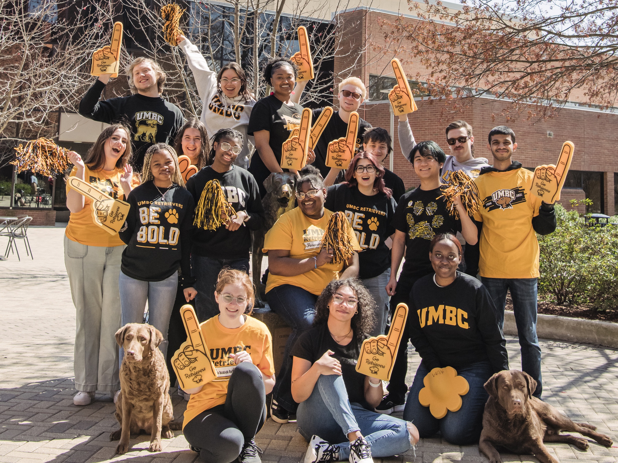 UMBC students dressed in black and gold gathered around the UMBC statue with a real dog on either side of the group.