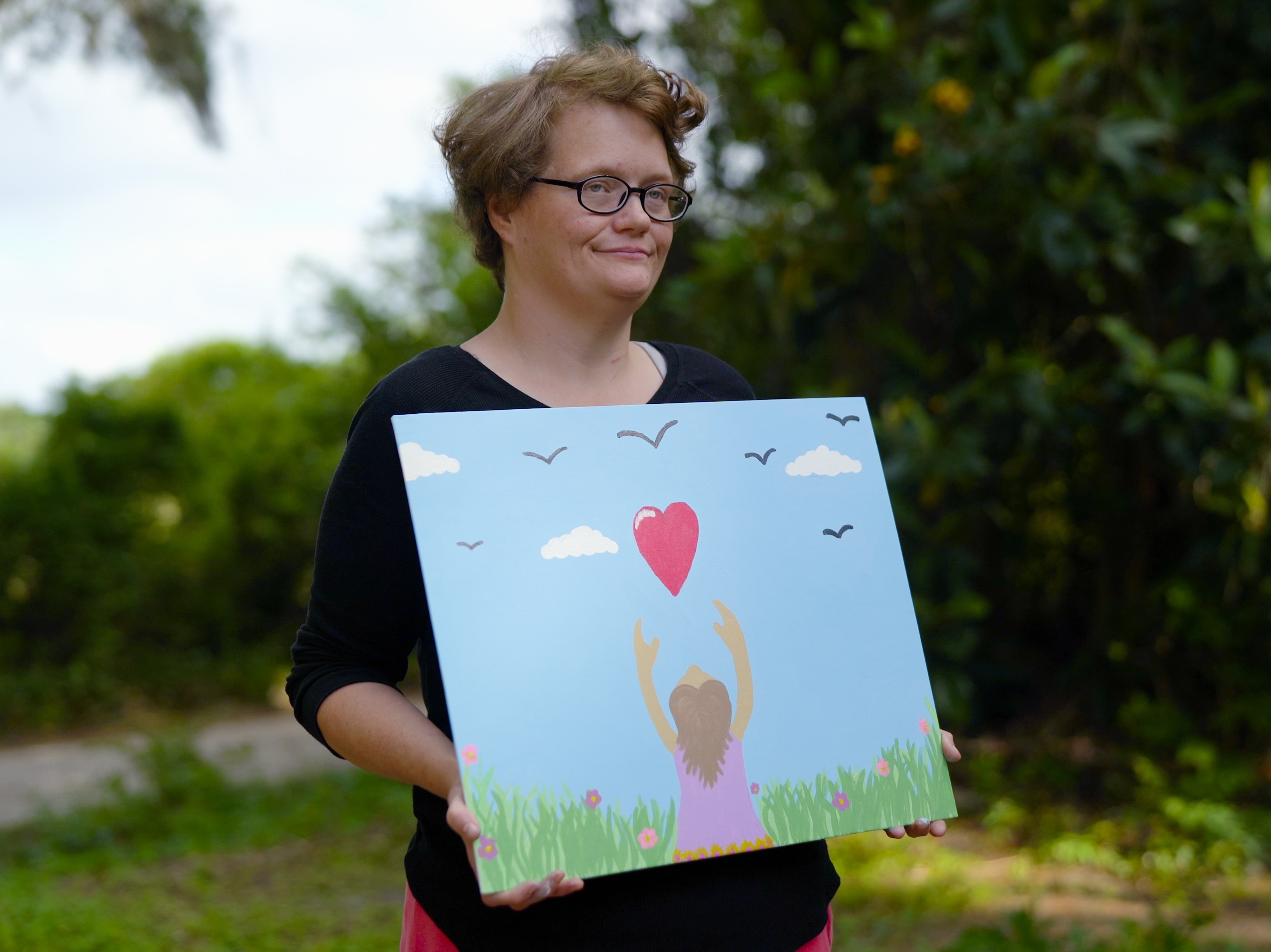 Michelle, a young white woman with glasses and short wavy light hair, holding her painting of a girl reaching for a heart in the sky