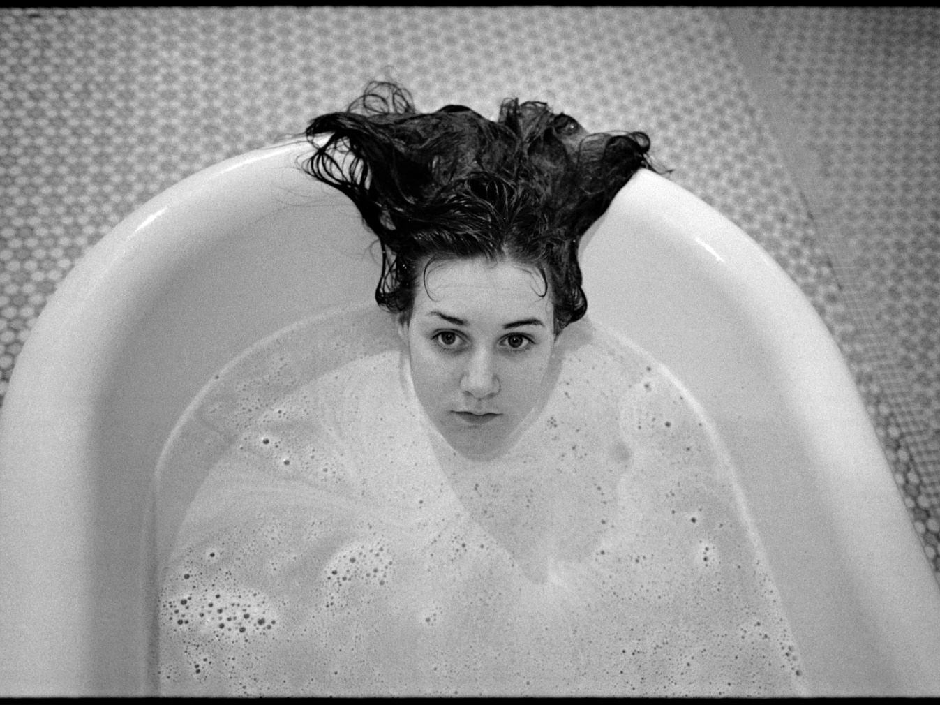 In a black and white photo, a woman with dark hair is submerged in a bathtub, with only her head visible.